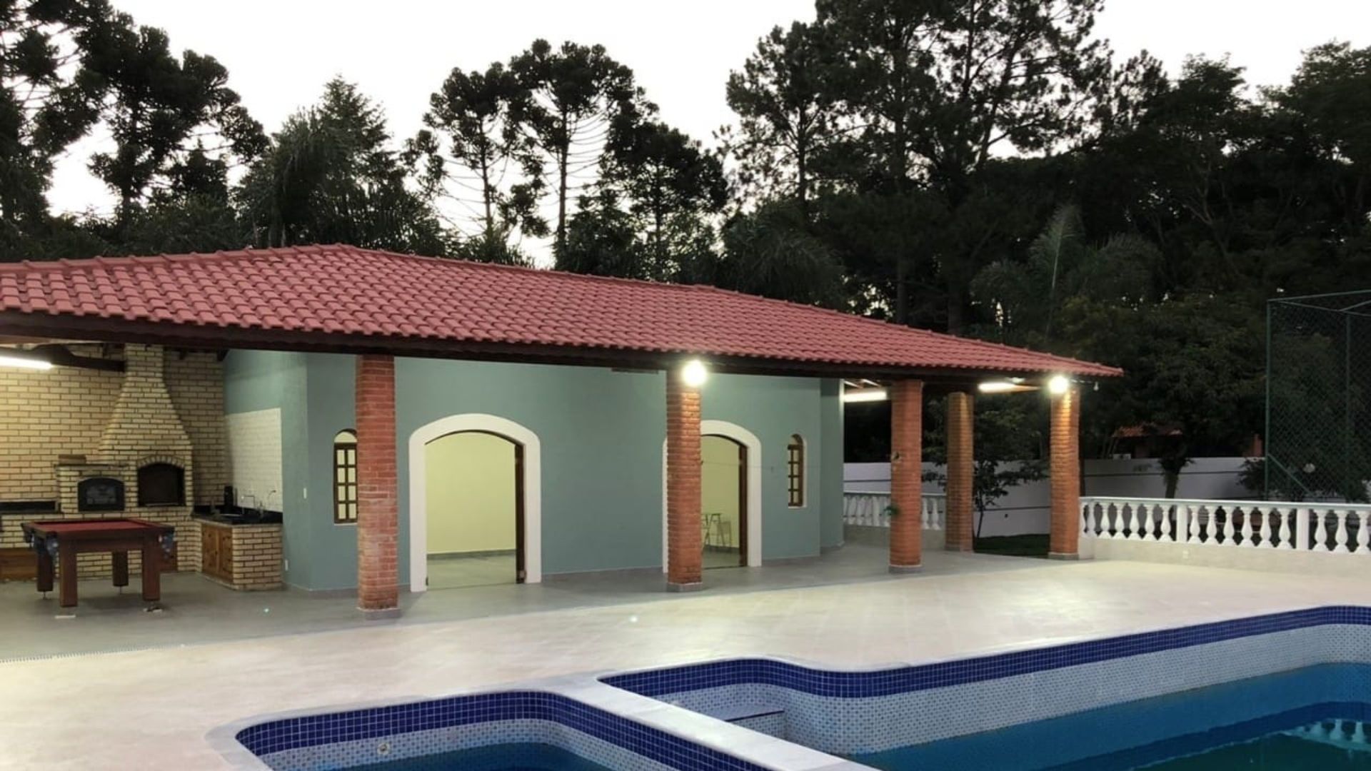 Área de lazer de casa de alto padrão reformada em São Paulo, piscina com borda em azulejo azul e branco, cobertura com telhado colonial e colunas em tijolo aparente, área gourmet com churrasqueira, jardim arborizado ao fundo, fotografia realista ao entardecer