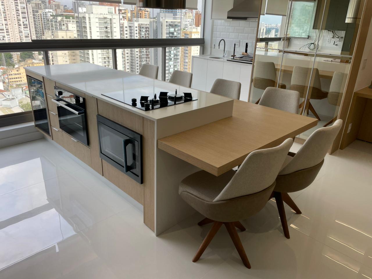 Cozinha planejada de alto padrão reformada em apartamento com vista para a cidade de São Paulo, ilha central em mármore com cooktop e forno embutido, mesa de jantar em madeira integrada, cadeiras estofadas, fotografia realista com luz natural
